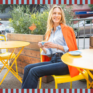 Woman wearing dark denim jeans sitting at an outdoor café holding a coffee cup