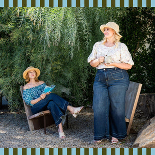 Two women wearing denim in a garden, one sitting and reading a book, the other standing and holding a mug.