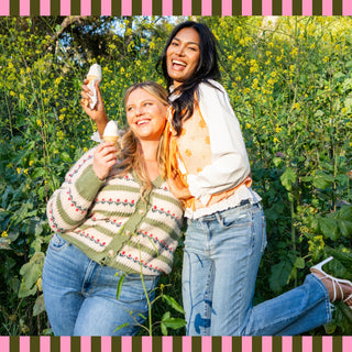 Two women smiling and holding vanilla soft-serve ice cream cones while posing in a sunlit field of yellow wildflowers, framed by a pink and olive striped border.